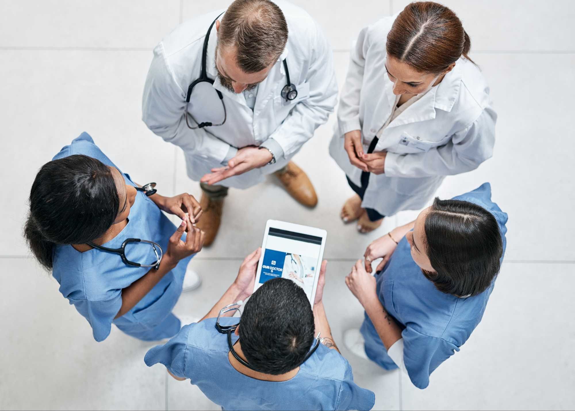 An overhead view of a medical team in scrubs and white coats engaged in a collaborative discussion around a tablet, highlighting teamwork in a clinical setting.