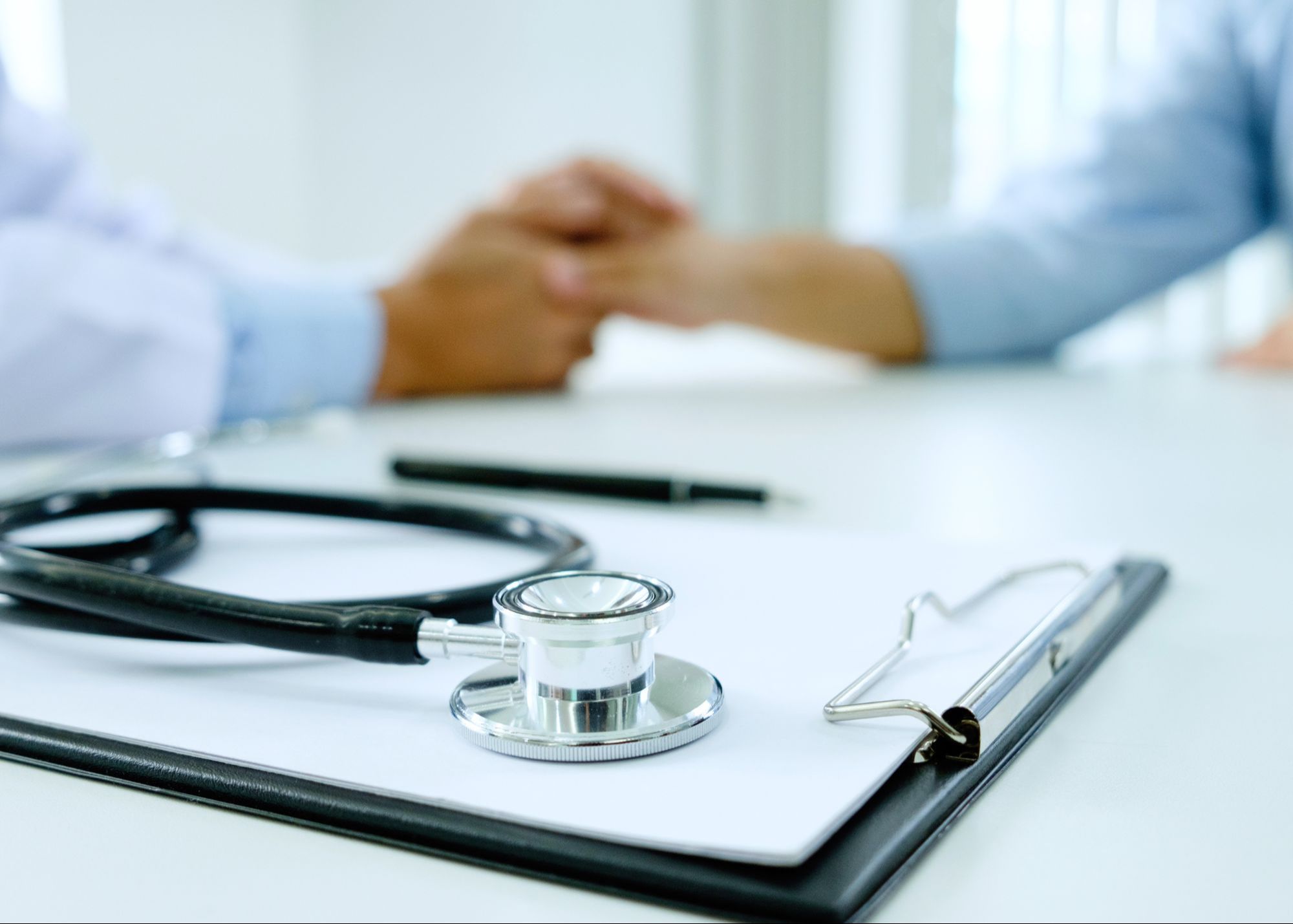 A doctor's desk with a stethoscope and clipboard, foregrounding a comforting hand gesture to a patient during a consultation.