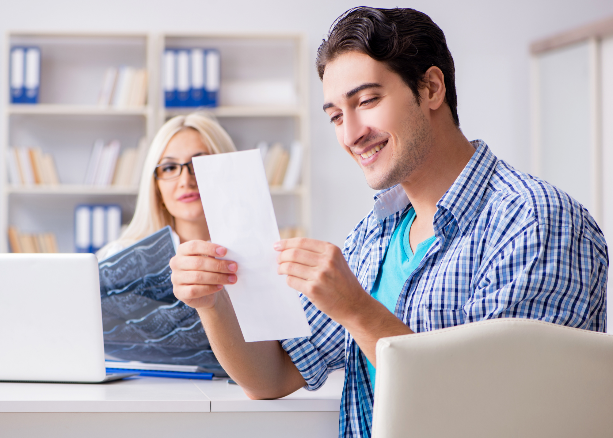 A cheerful man reading a document with a pleased expression as a female physician with glasses observes from behind a laptop in a medical office setting.